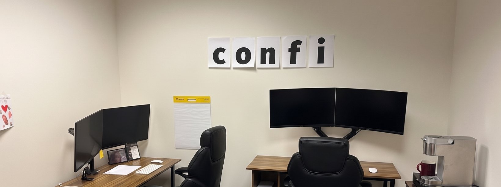 A small office: two desks, two chairs, a blank flipchart, a coffee machine, and the word confi printed on five sheets of paper taped to the wall